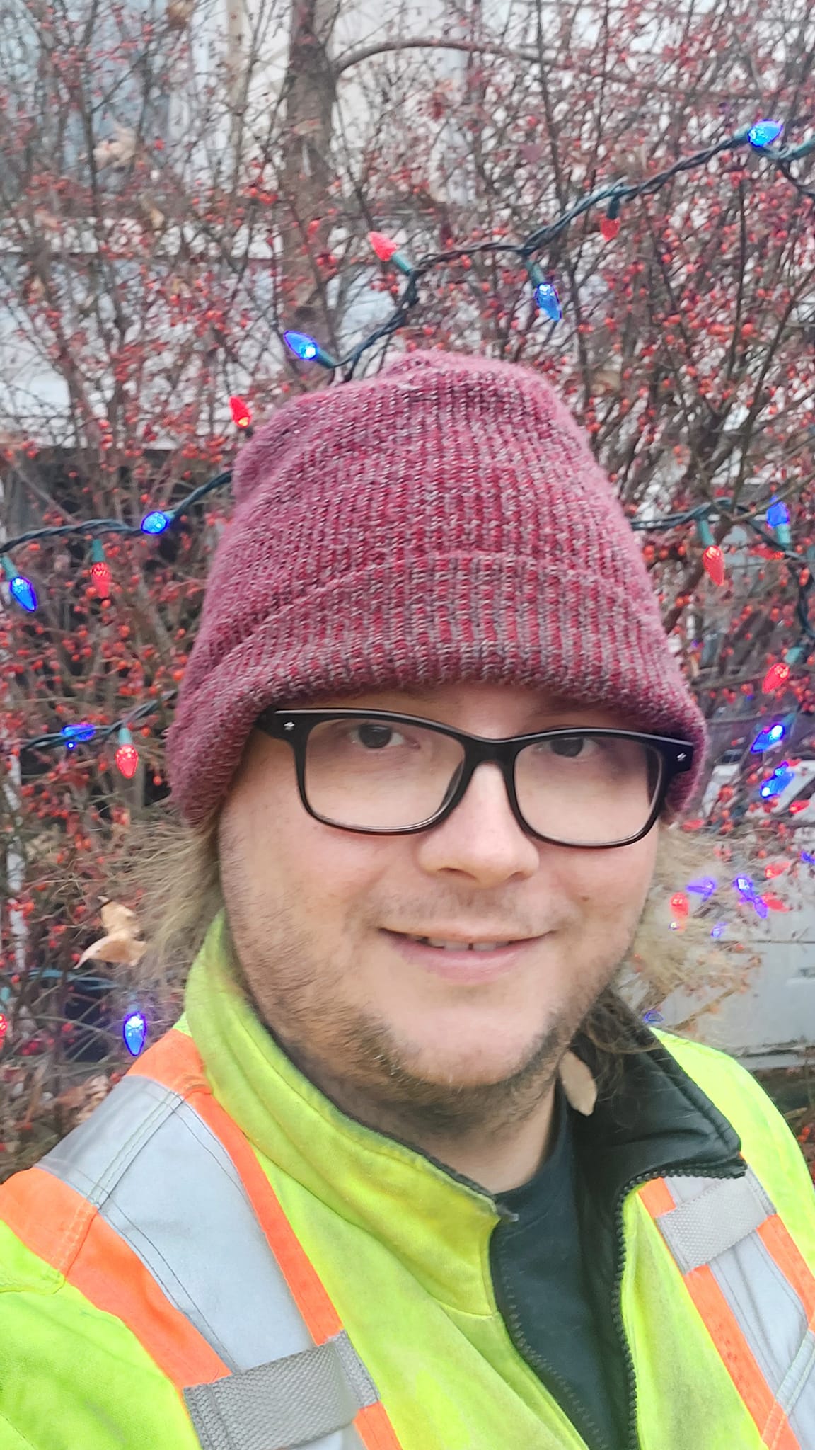 A man with long green hair wears a red toque and high visibility winter coat stands in front of a bush with red and blue string lights on it